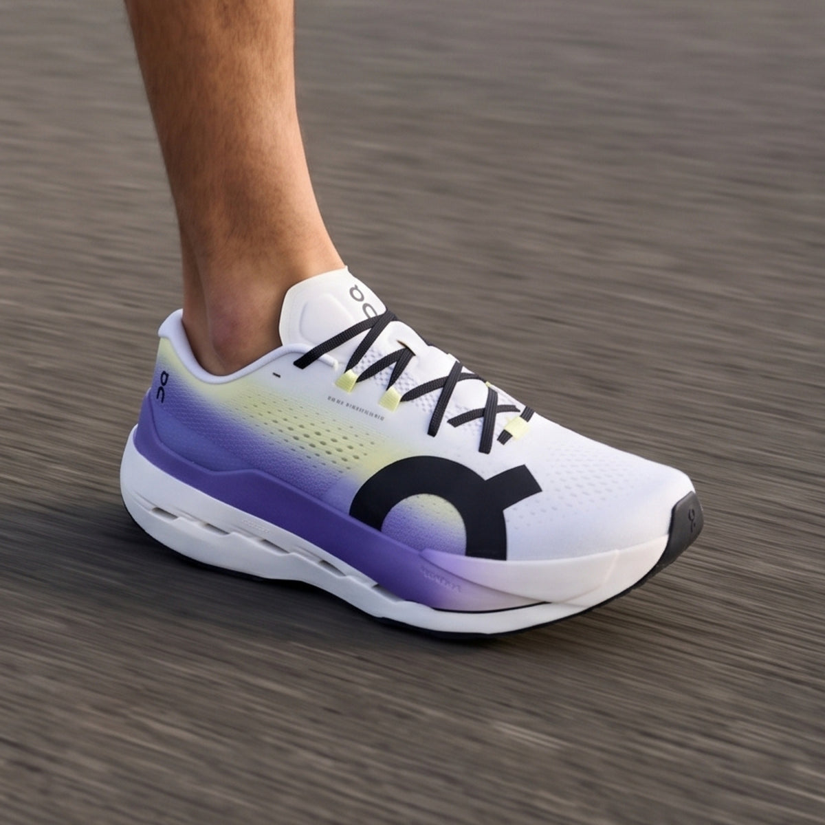 A close-up of a person's foot wearing an On running shoe, captured in motion on a blurred gray asphalt surface. The shoe is white with a purple and yellow gradient on the midsole and collar, featuring a large black On logo on the side and black laces.