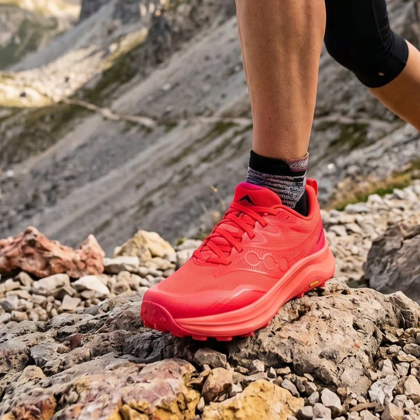 Person wearing a red Saucony running shoe on rocky terrain with mountains in the background