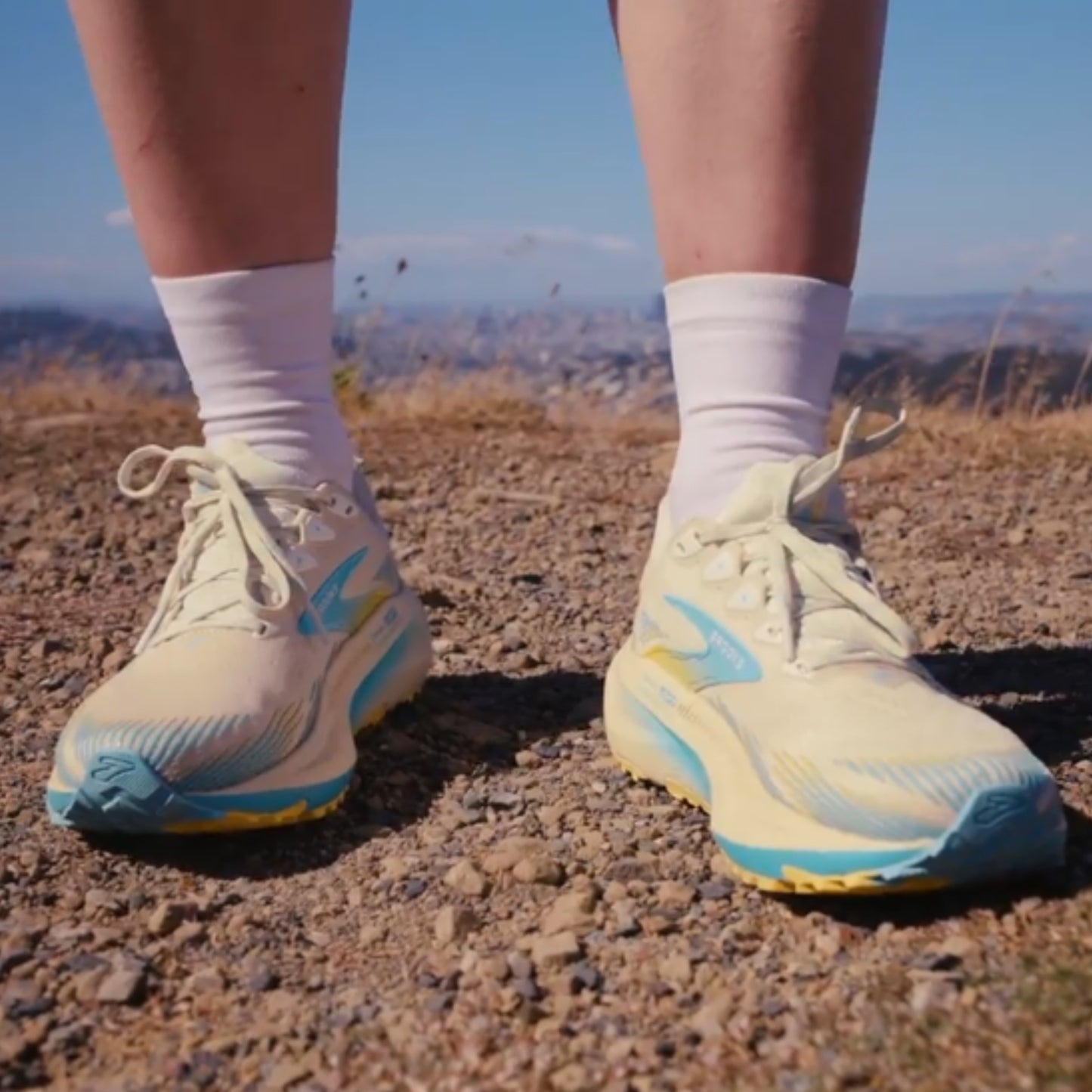 Beige and blue Brooks running shoes on a person's feet with a mountainous landscape in the background.