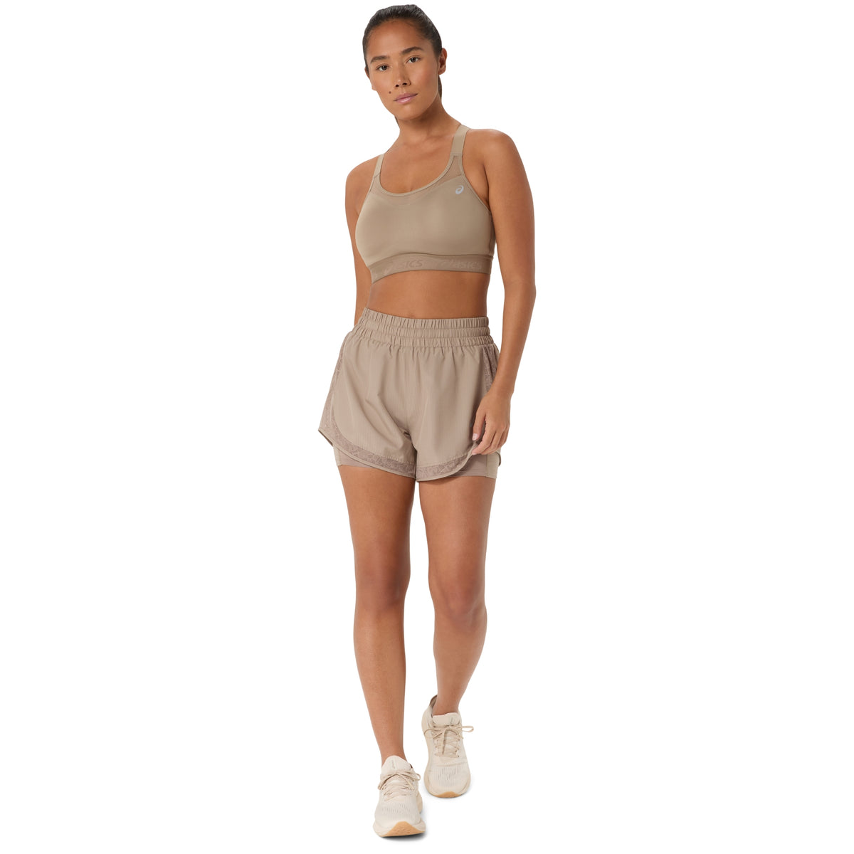 A woman modeling a matching beige athletic set consisting of a sports bra and shorts, paired with light beige sneakers, against a solid white background.