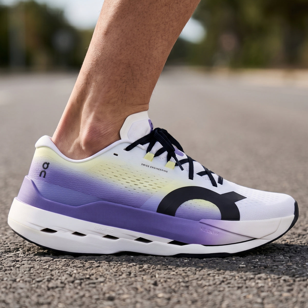 A close-up of a person's foot wearing an On running shoe on an asphalt surface. The shoe features a white upper with a purple and yellow gradient, black laces, and a black On logo.