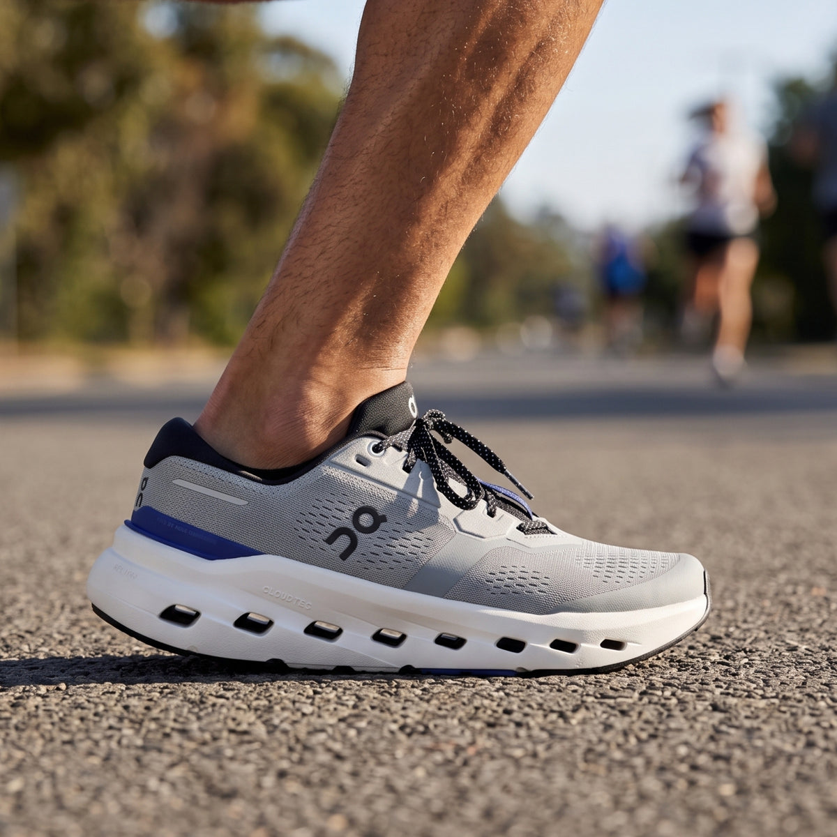 A close-up of a runner's foot wearing a light gray On running shoe with a white sole, black laces, and a blue accent stripe, standing on an asphalt road with blurred runners in the background.