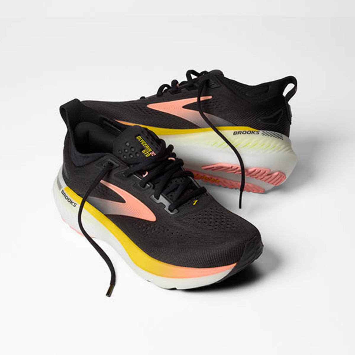 A pair of Brooks running shoes in black with coral pink, yellow, and white accents, displayed on a clean light gray background.
