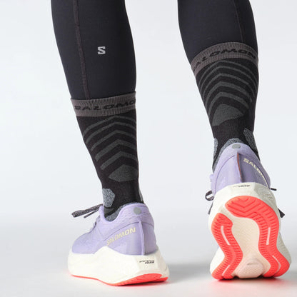 Person wearing black compression socks and purple Salomon running shoes with red soles on a light gray background