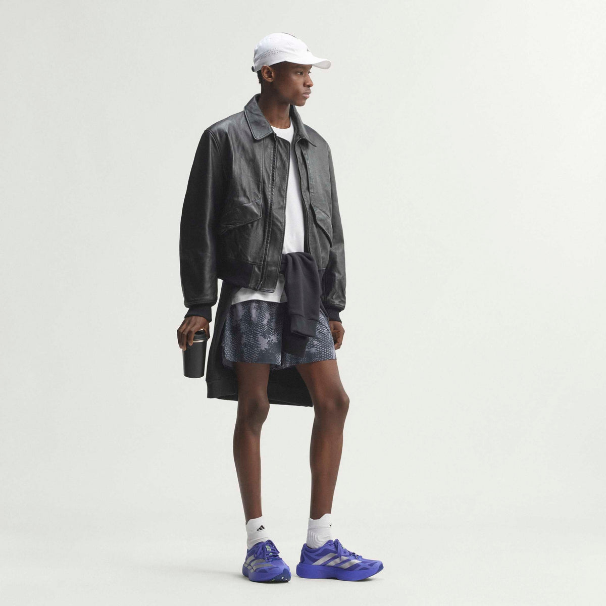 A model stands in profile wearing a black leather jacket, a white t-shirt, a patterned denim skirt, a white cap, white socks, and blue sneakers, against a plain light gray studio background.