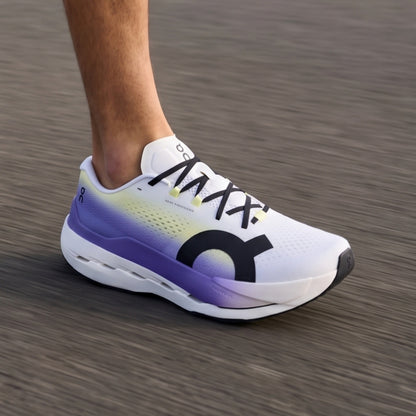 A close-up of a person's foot wearing an On running shoe, captured in motion on a blurred gray asphalt surface. The shoe is white with a purple and yellow gradient on the midsole and collar, featuring a large black On logo on the side and black laces.