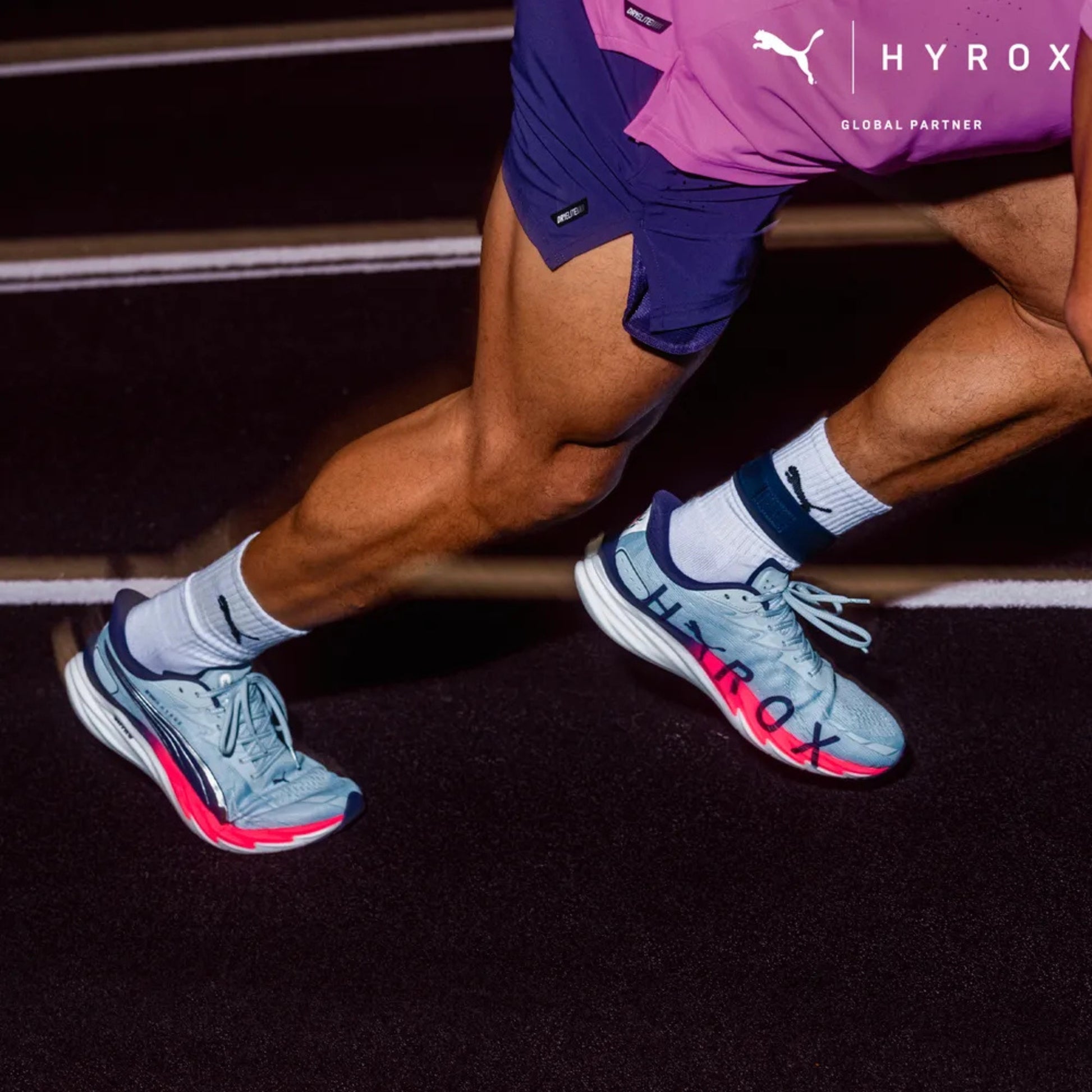 Person wearing light Puma shoes running on a track wearing blue running shoes with 'Hyrox' branding.