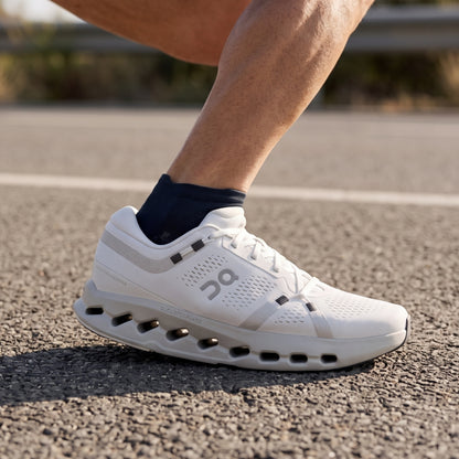 A close-up of a runner's foot wearing a white On running shoe with a dark navy sock, standing on an asphalt road.