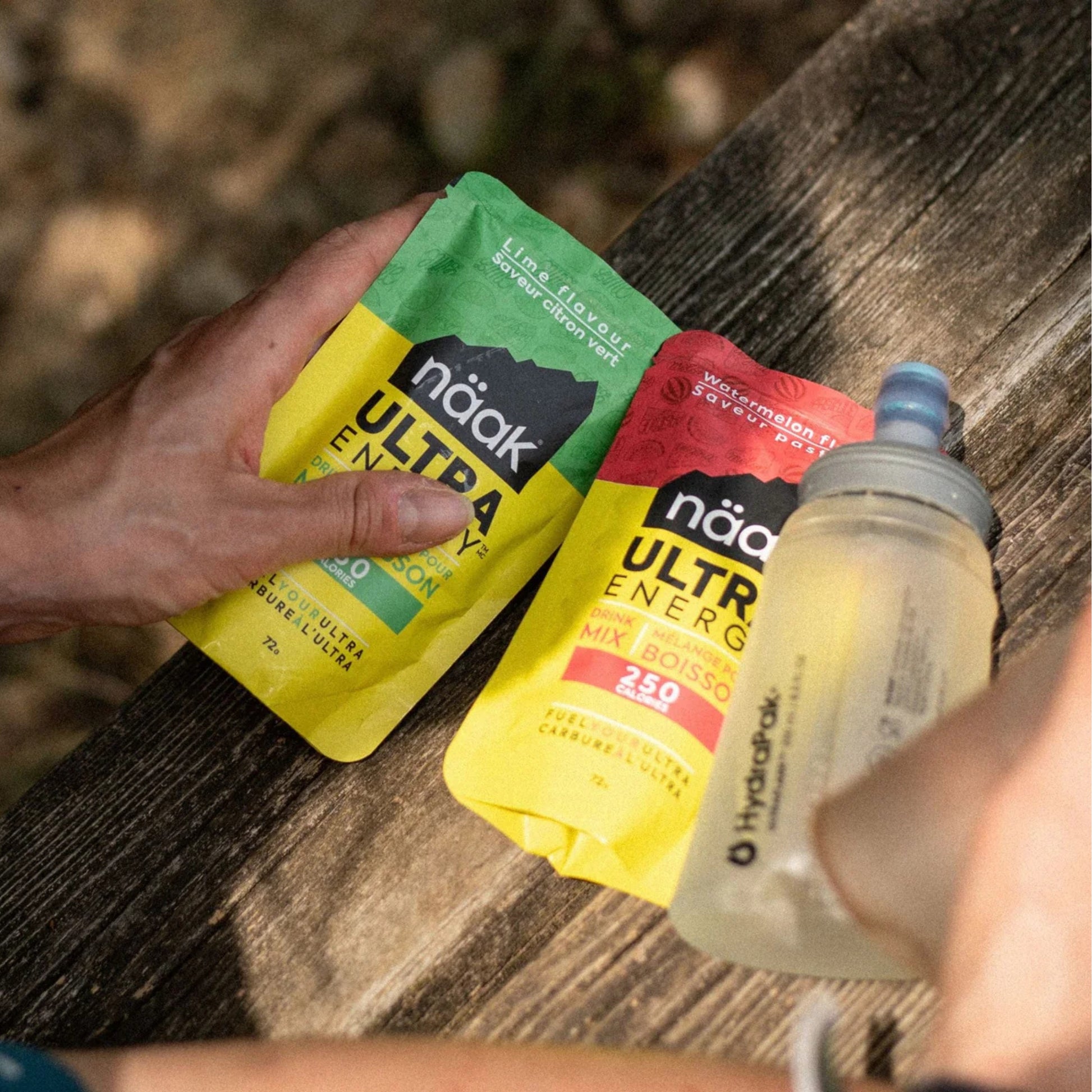 Hand holding two náak Ultra Energy Mix packets and a water bottle on a wooden surface.