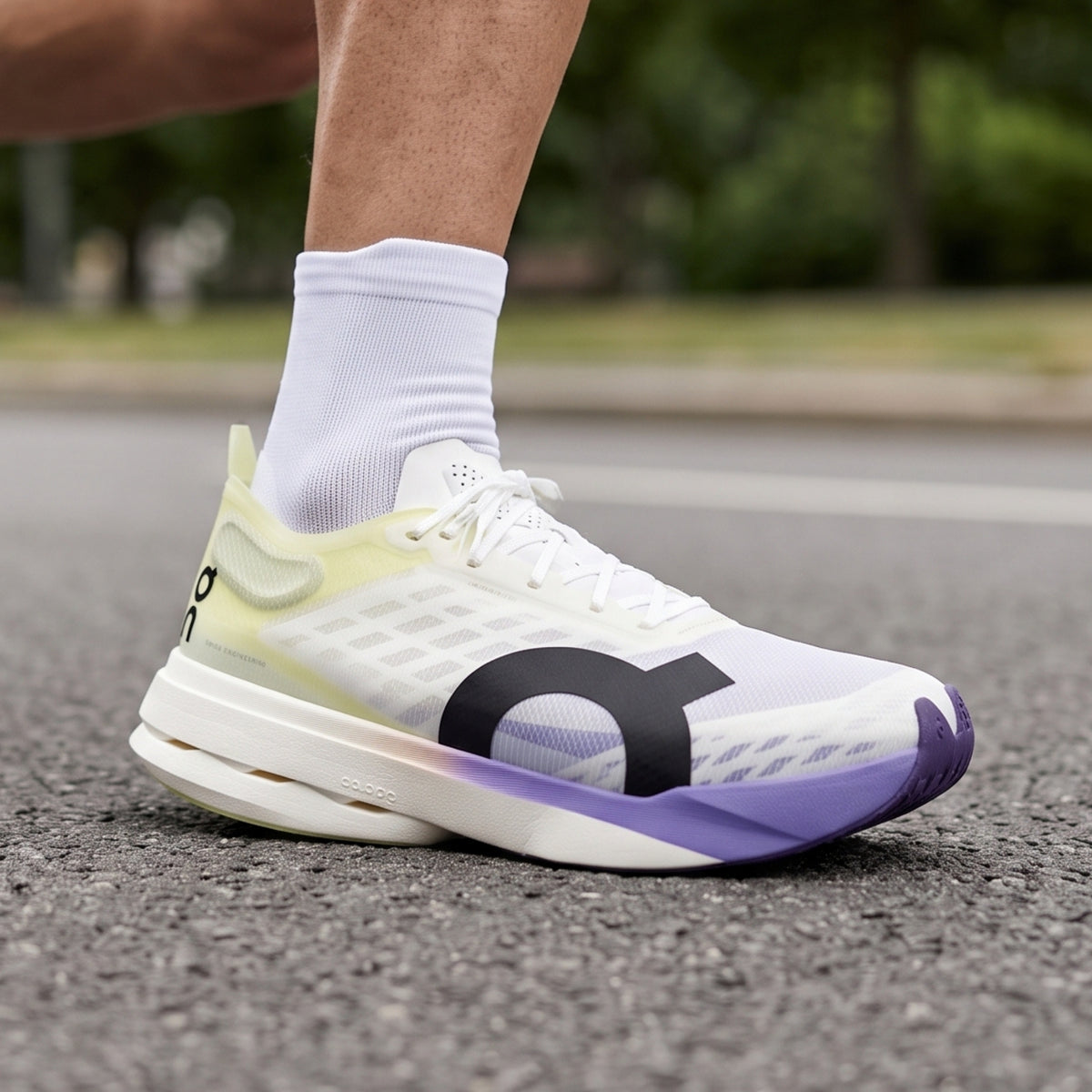 A close-up of a person's foot wearing a white On running shoe with a yellow-green heel, purple midsole accent, and a large black On logo on the side, standing on asphalt outdoors.