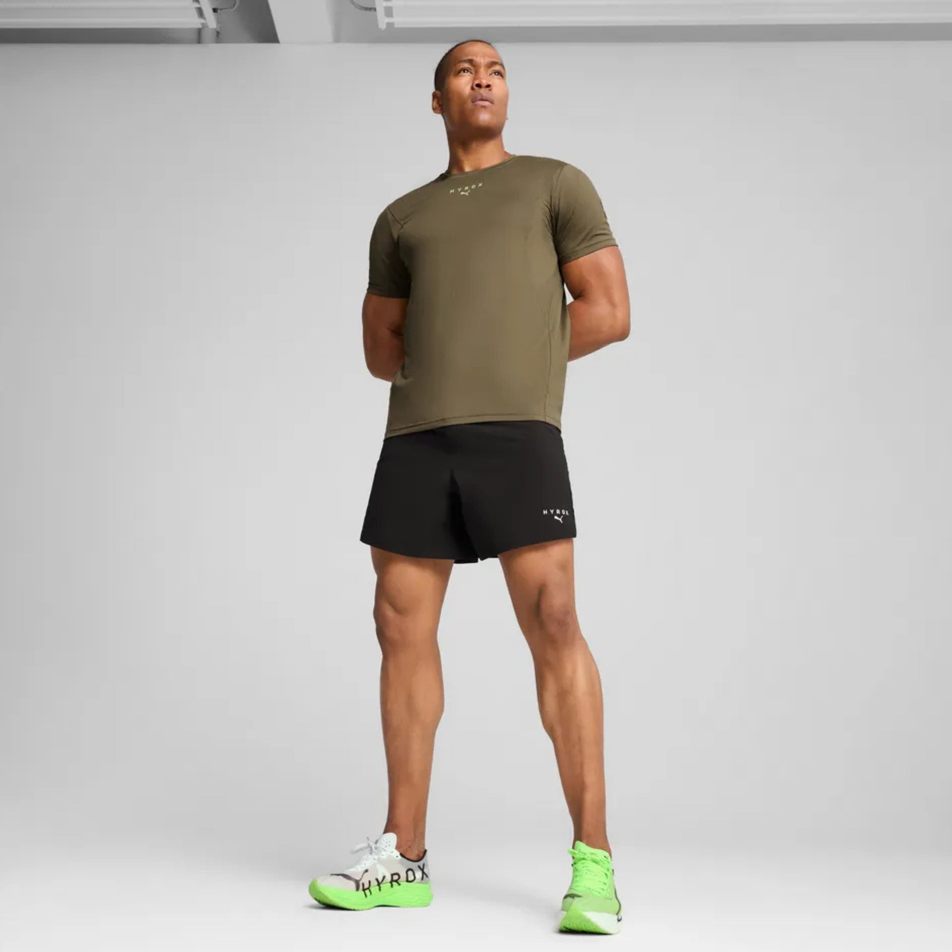 Man wearing an olive green t-shirt and black shorts with neon green shoes on a gray background