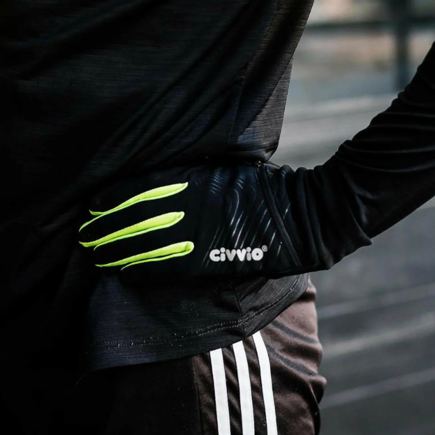 Person wearing a black jacket with black gloves with green accents and 'civvio' branding, standing by a body of water.