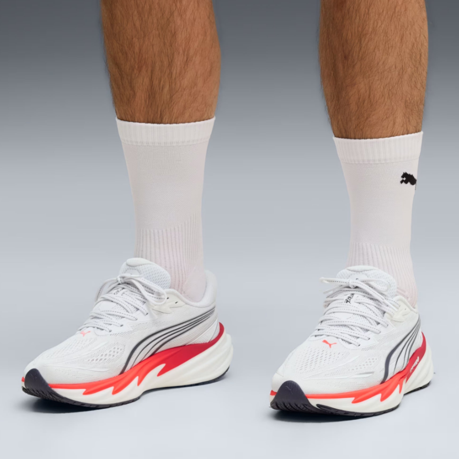 White Puma running shoes with red and gray accents worn with white crew socks, shown on a model's feet against a gray studio background.