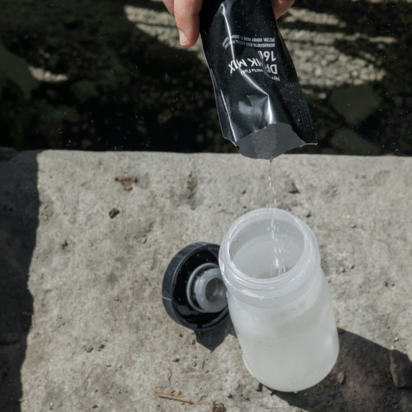 Person pouring a clear liquid from a black container into a white container on a concrete surface.