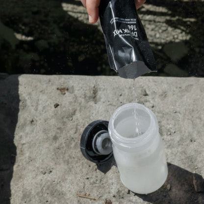 Person pouring a clear liquid from a black container into a white container on a concrete surface.