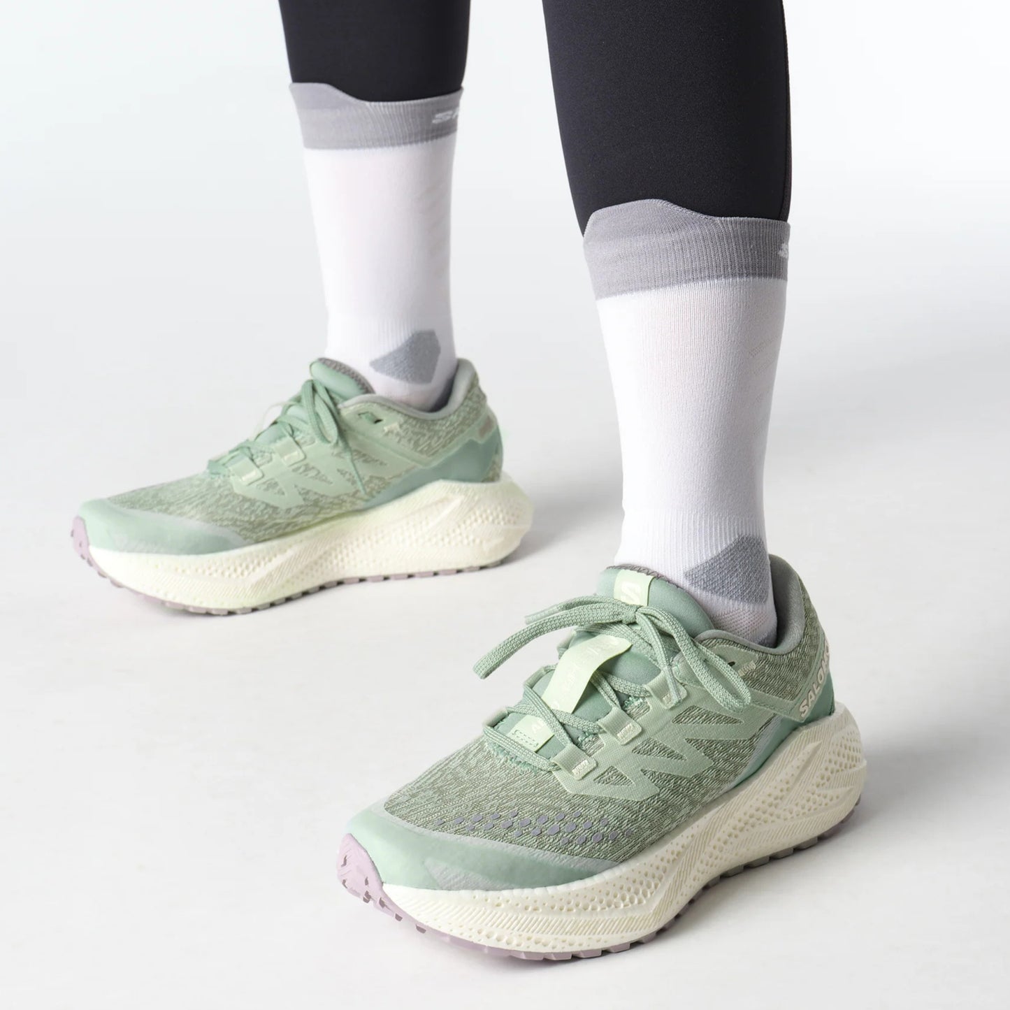 Light green Salomon running shoes worn with dark leggings and white socks on a light gray background