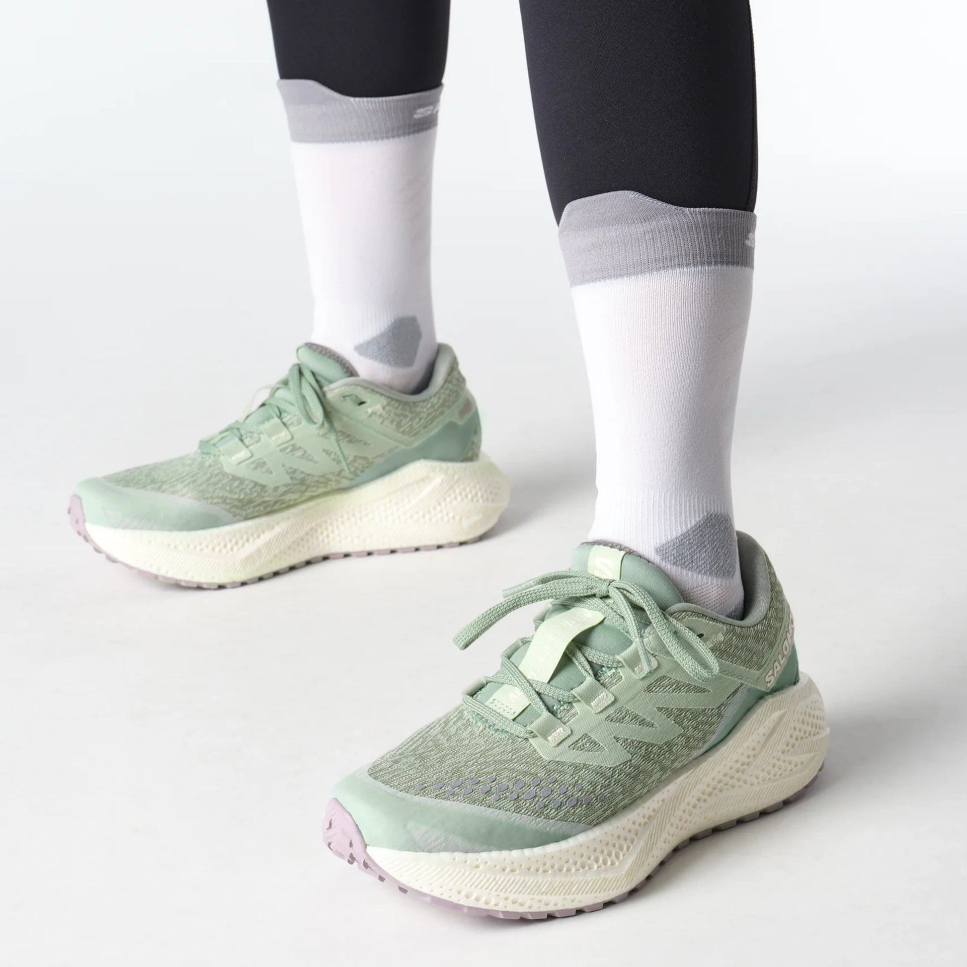 Light green Salomon running shoes worn with dark leggings and white socks on a light gray background