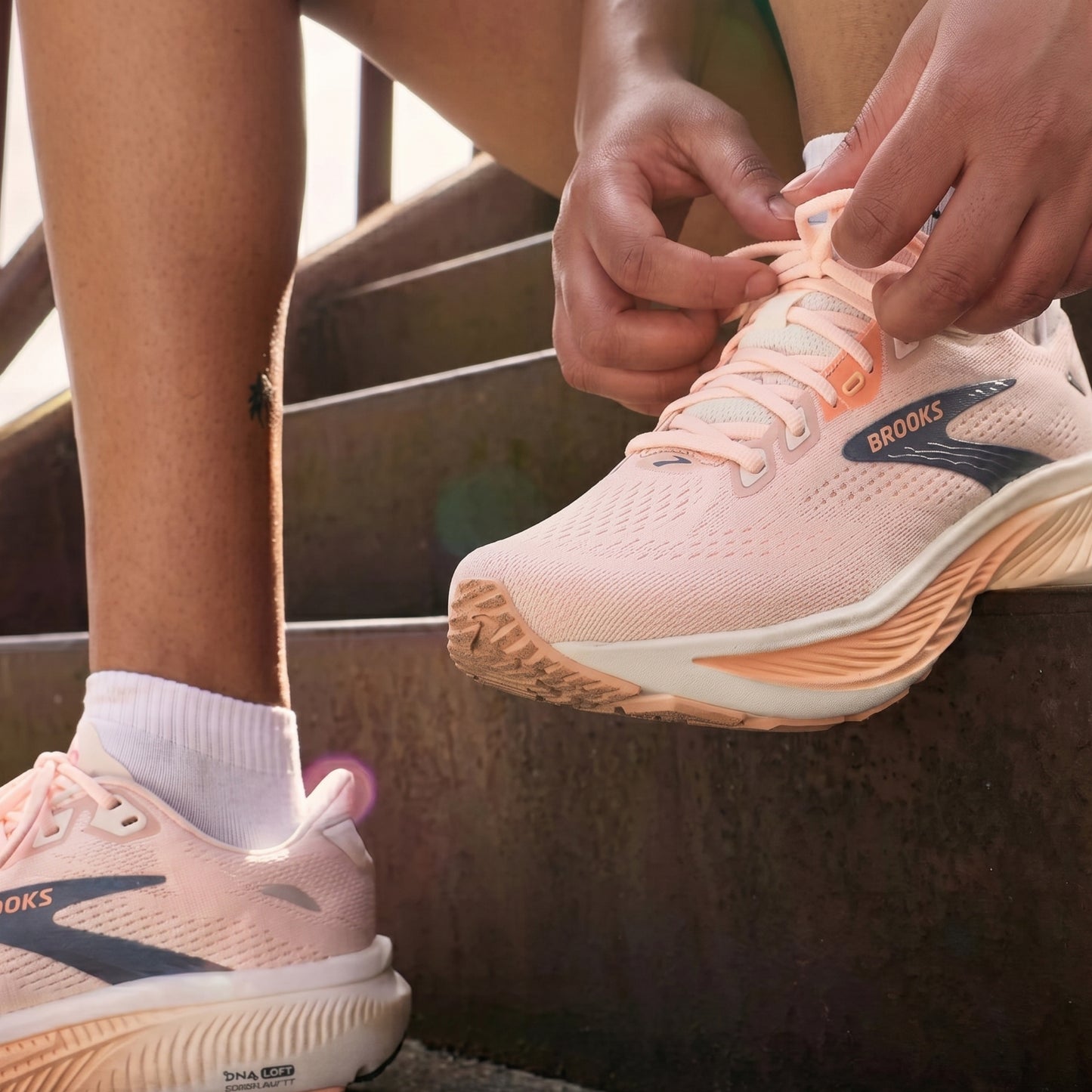 A close-up lifestyle shot of a person sitting on outdoor steps, tying the laces of a light pink Brooks running shoe while wearing the same shoe on their foot.