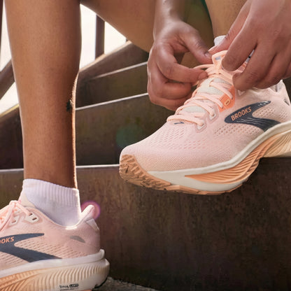 A close-up lifestyle shot of a person sitting on outdoor steps, tying the laces of a light pink Brooks running shoe while wearing the same shoe on their foot.