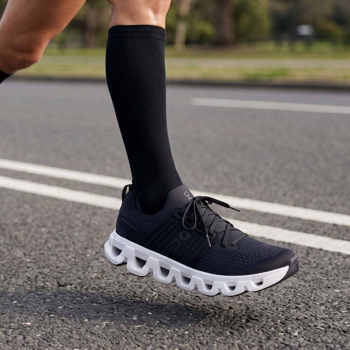 A close-up of a runner's lower leg wearing a black On running shoe with a white cloud-like sole and a black compression sock, captured mid-stride on an asphalt road.