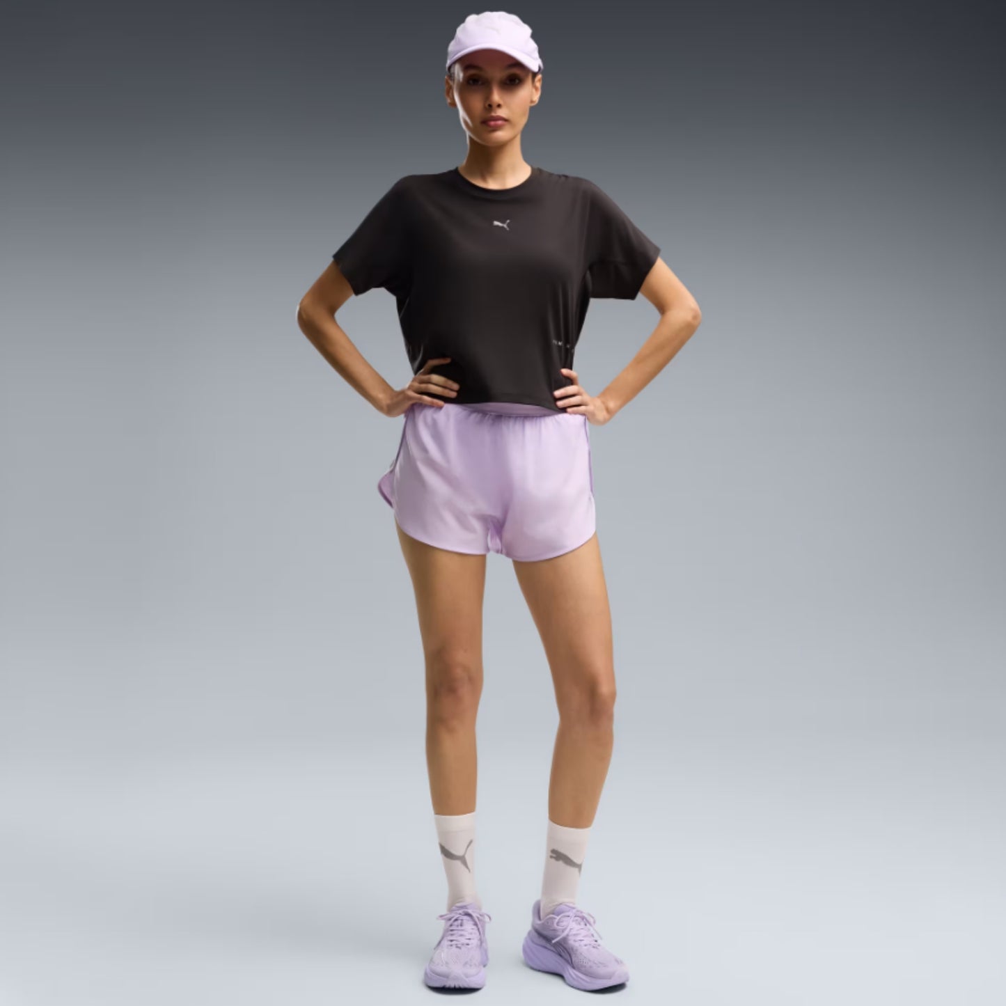 A woman modeling athletic wear in a studio setting, wearing a black short-sleeve athletic top, light lavender shorts, a white cap, white socks, and light purple sneakers against a gray gradient background.