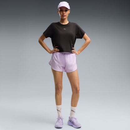 A woman modeling athletic wear in a studio setting, wearing a black short-sleeve athletic top, light lavender shorts, a white cap, white socks, and light purple sneakers against a gray gradient background.
