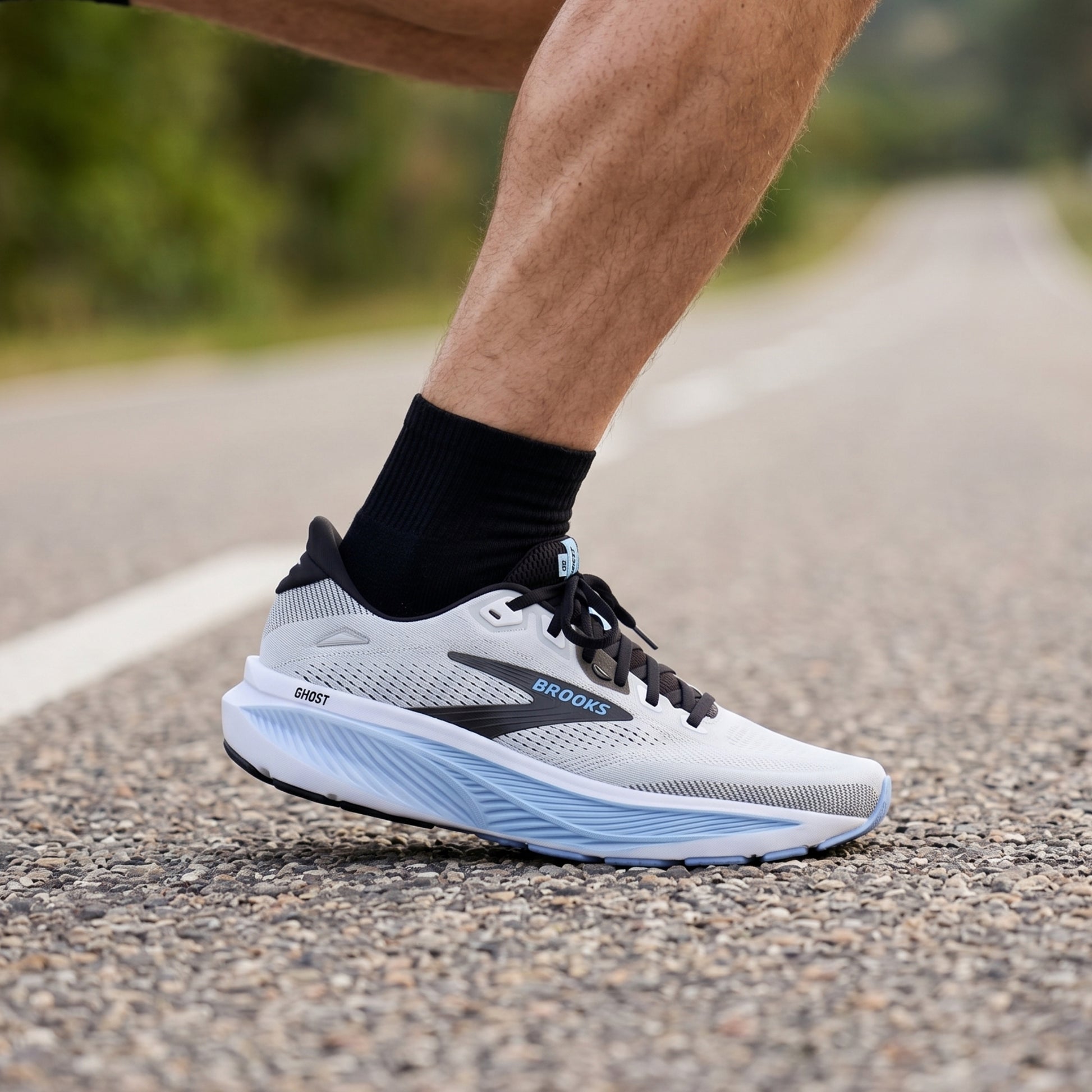 A close-up of a runner's foot wearing a Brooks Ghost running shoe on an asphalt road with a blurred natural background.