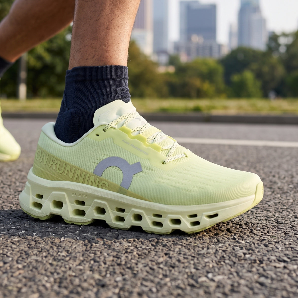 A close-up of a person's foot wearing a light yellow-green ON Running shoe with a matching thick cushioned sole, paired with a dark navy sock, set on an asphalt road with a blurred city skyline in the background.