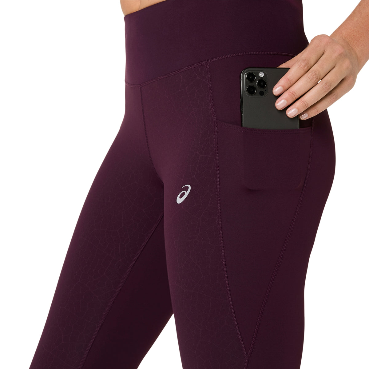 Close-up of a person wearing high-waisted athletic leggings in deep plum purple, showing a side pocket holding a black smartphone. The ASICS logo is visible on the thigh.