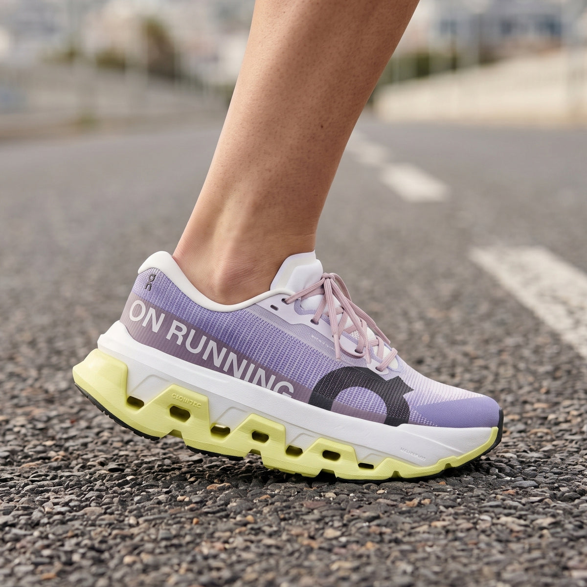 A close-up of a person's leg wearing a purple and white On running shoe with a chunky yellow-green sole, standing on an asphalt road.
