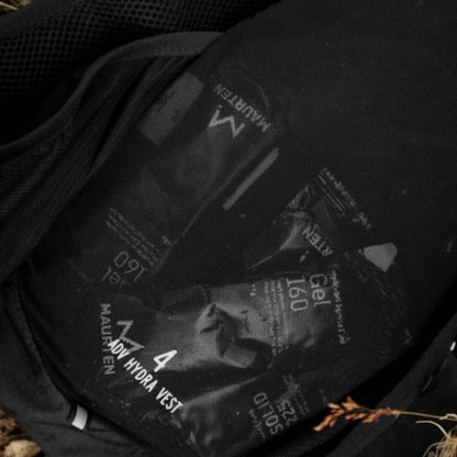 A close-up of a black hydration vest or backpack interior showing several black hydration packet sachets printed with white text, including 'MAURTEN GEL'