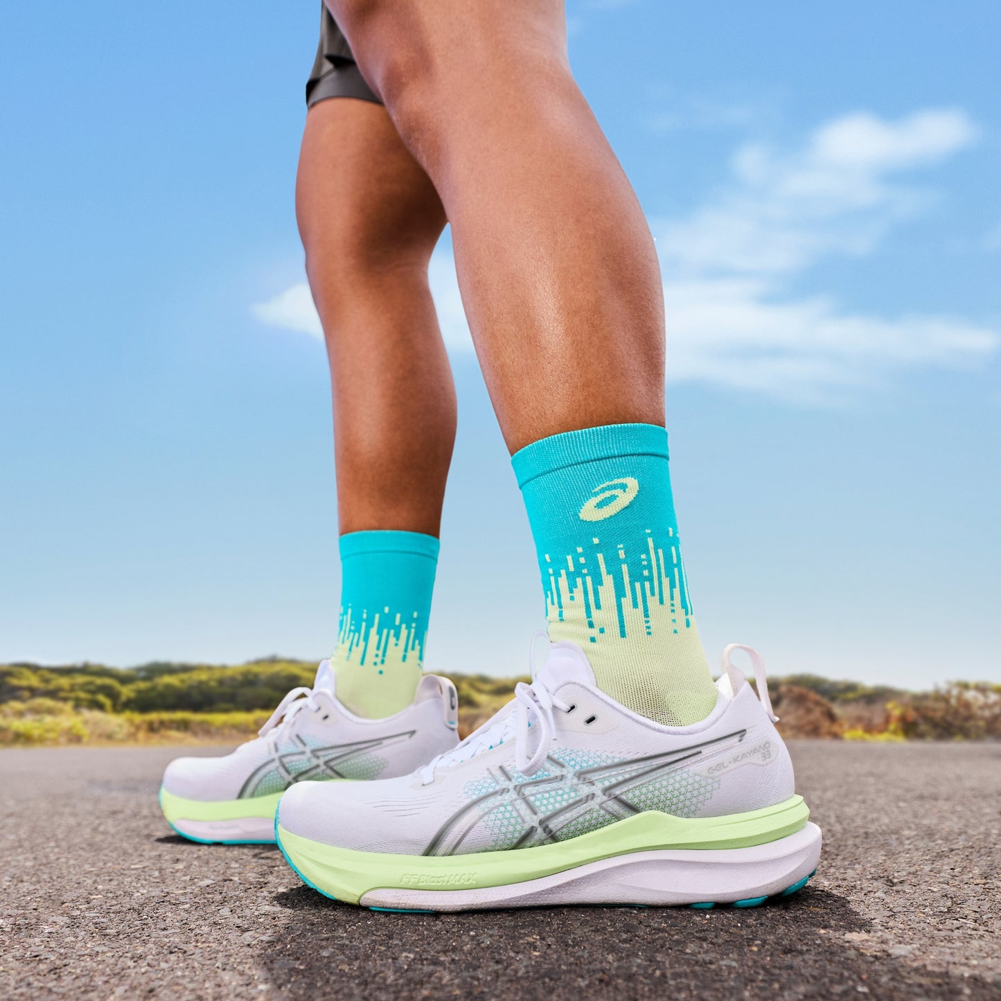 Person wearing turquoise socks with a brand logo and white running shoes on a road with a blue sky background.