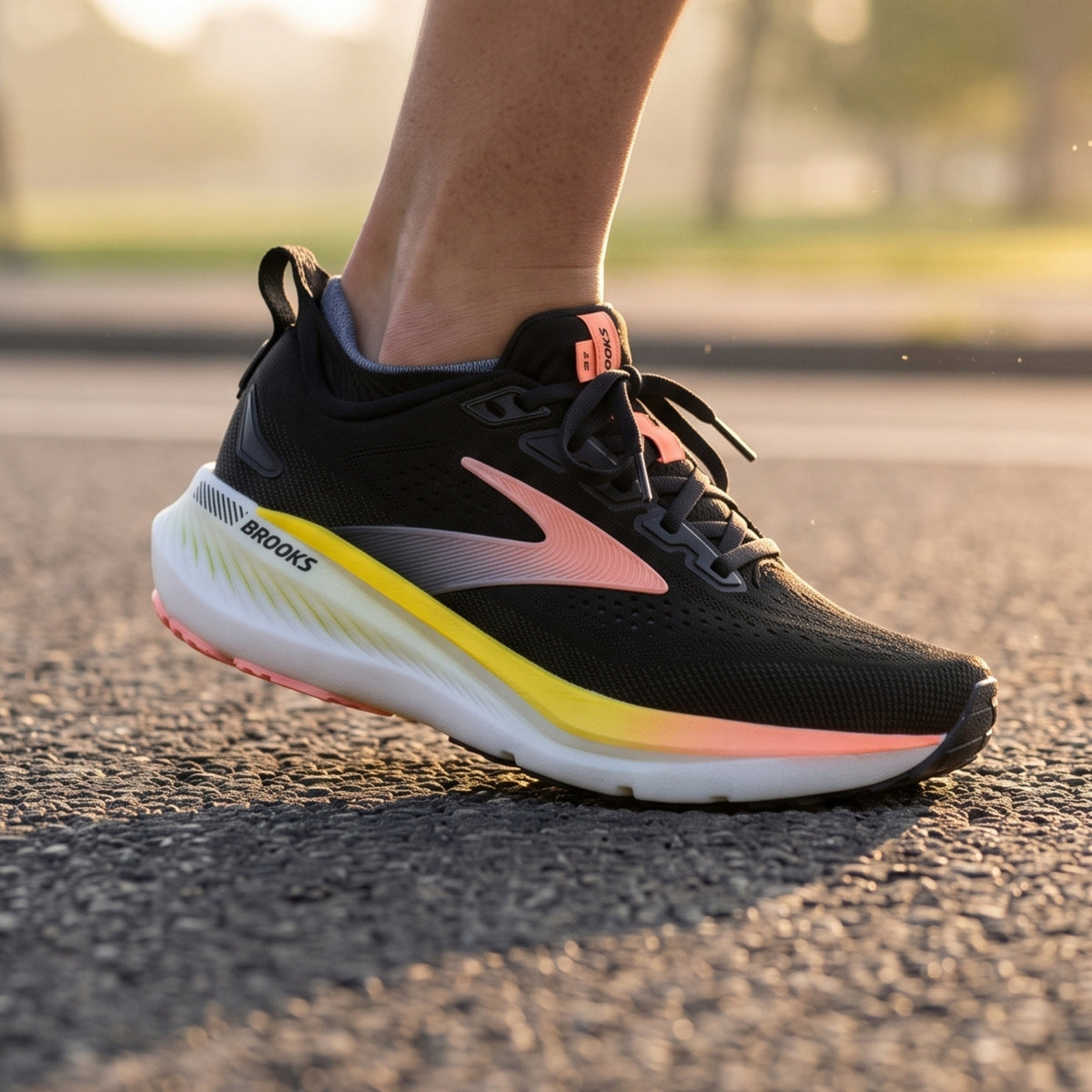 A close-up of a Brooks running shoe being worn, with a person's lower leg visible. The shoe is black with a pink and yellow gradient logo and midsole accent, photographed outdoors on asphalt with a blurred background.