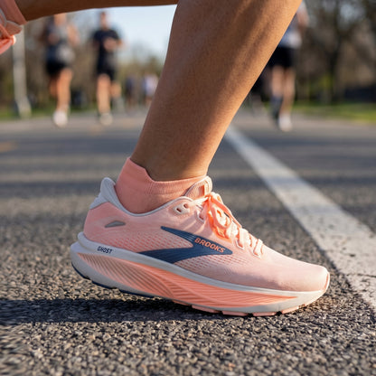 A close-up of a runner's foot wearing a Brooks Ghost running shoe in light pink with a dark blue logo and matching pink socks, standing on an asphalt running track with blurred runners in the background.