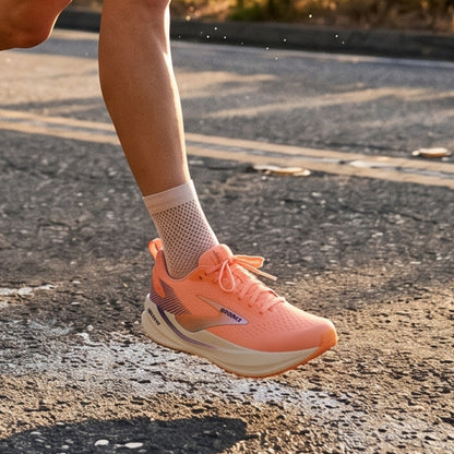 A close-up of a runner's foot wearing a coral pink Brooks running shoe and a white mesh sock, captured mid-jump on an asphalt road during warm golden-hour sunlight.