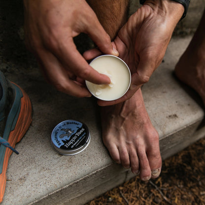 Person applying balm to their foot with a tin labeled 'Happie Toes' on a concrete surface.