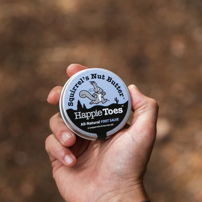 Hand holding a jar of 'Happie Toes' foot salve with a blurred natural background