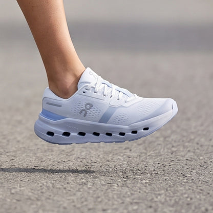 A white On running shoe with light blue accents, shown mid-air as a foot and lower leg approach it on a gray asphalt surface.