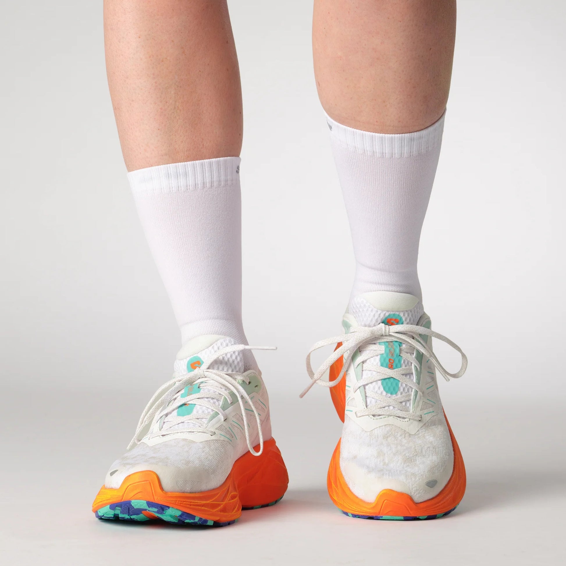 White running shoes with orange soles worn with white socks on a light gray background