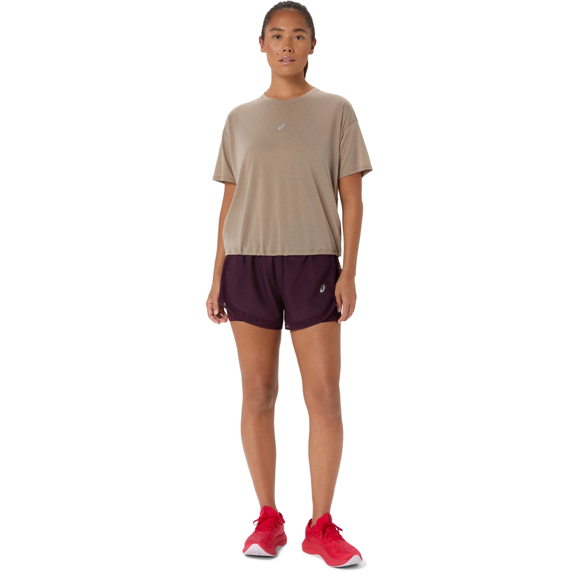 A woman modeling a light taupe short-sleeve athletic t-shirt and dark plum athletic shorts, paired with bright red running shoes, against a solid white background.