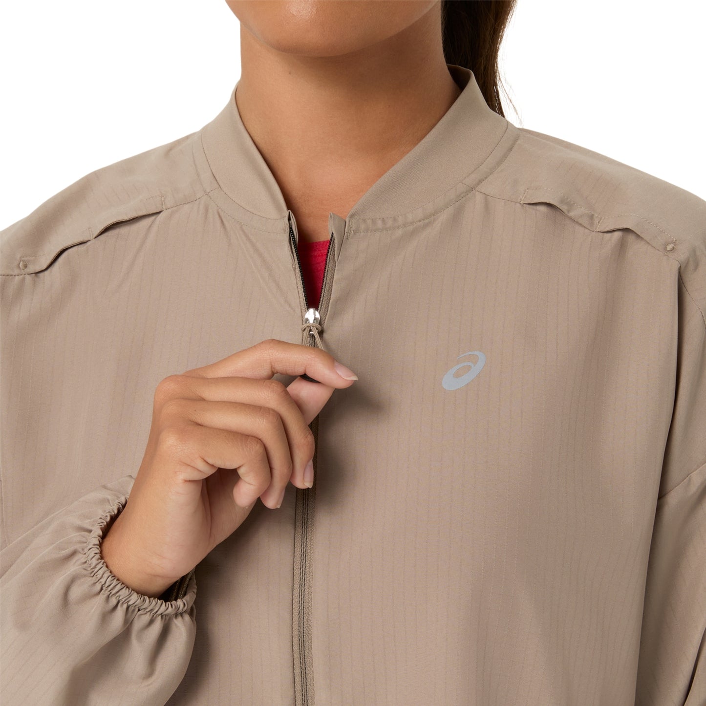 Close-up of a person wearing a beige lightweight zip-front jacket with a small light blue Asics logo on the chest, holding the zipper open against a white background.