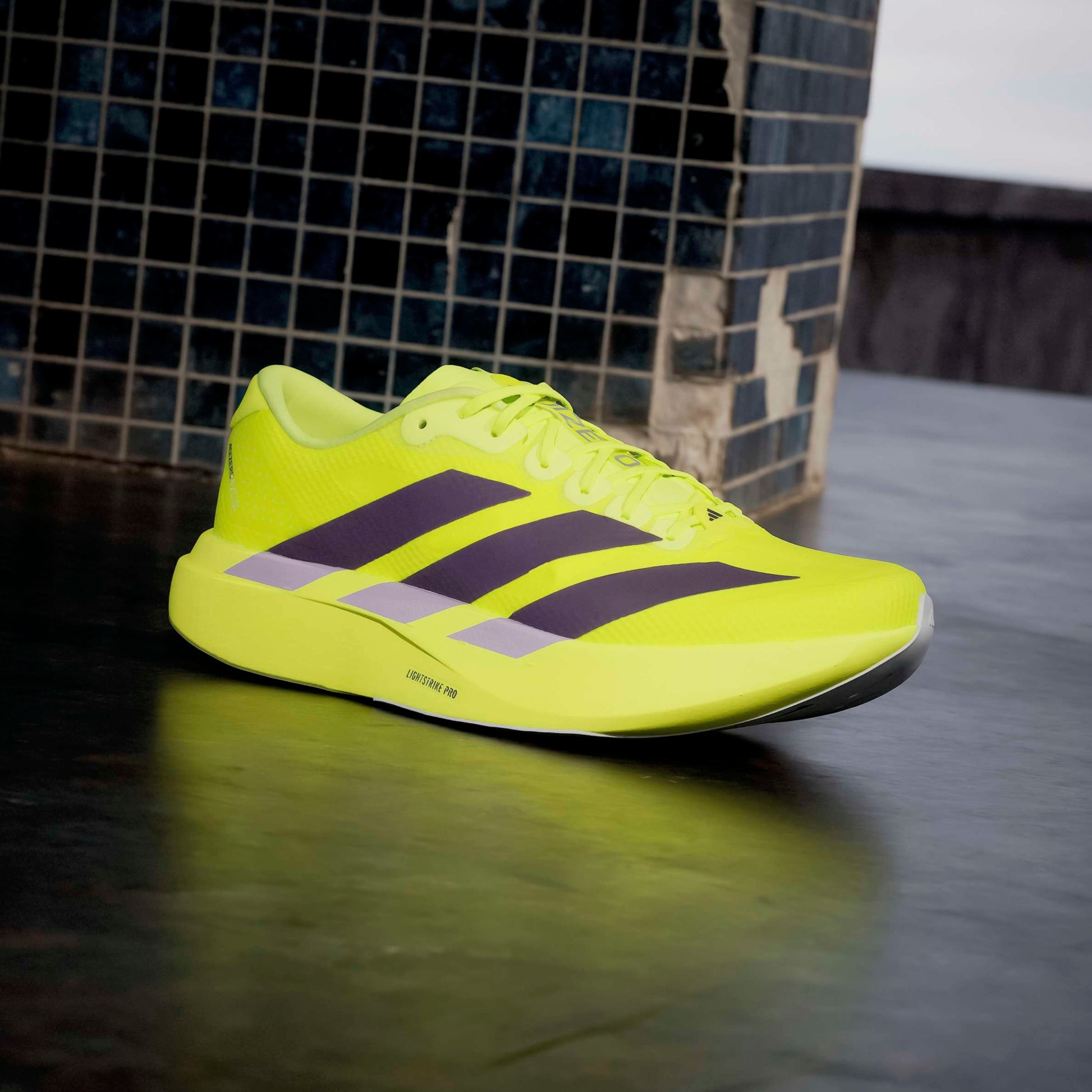 Bright yellow adidas running shoe with purple stripes on a reflective surface.