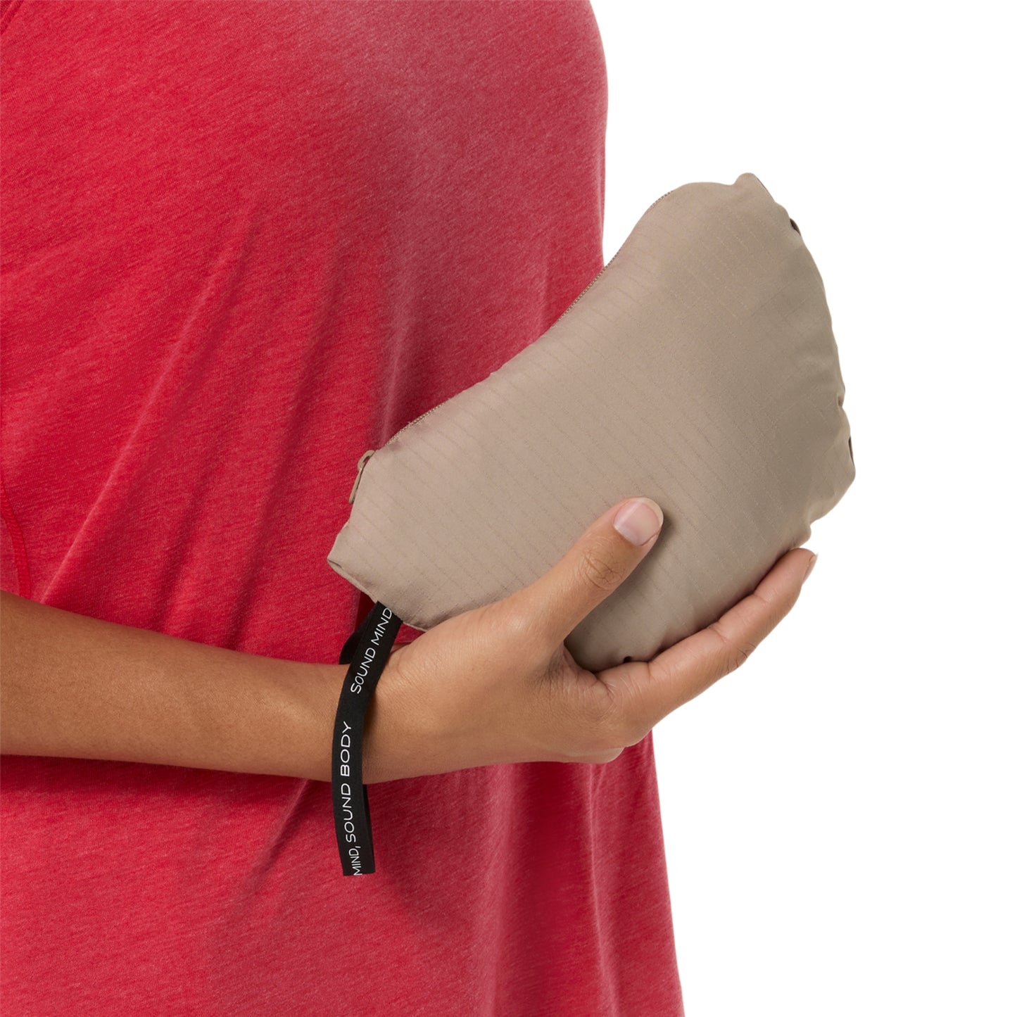 A person in a red shirt holds a compact, folded beige jacket bag against a white background. A black fabric tag with white text reading 'THE SNOOZY BODY' is visible near the bottom of the item.