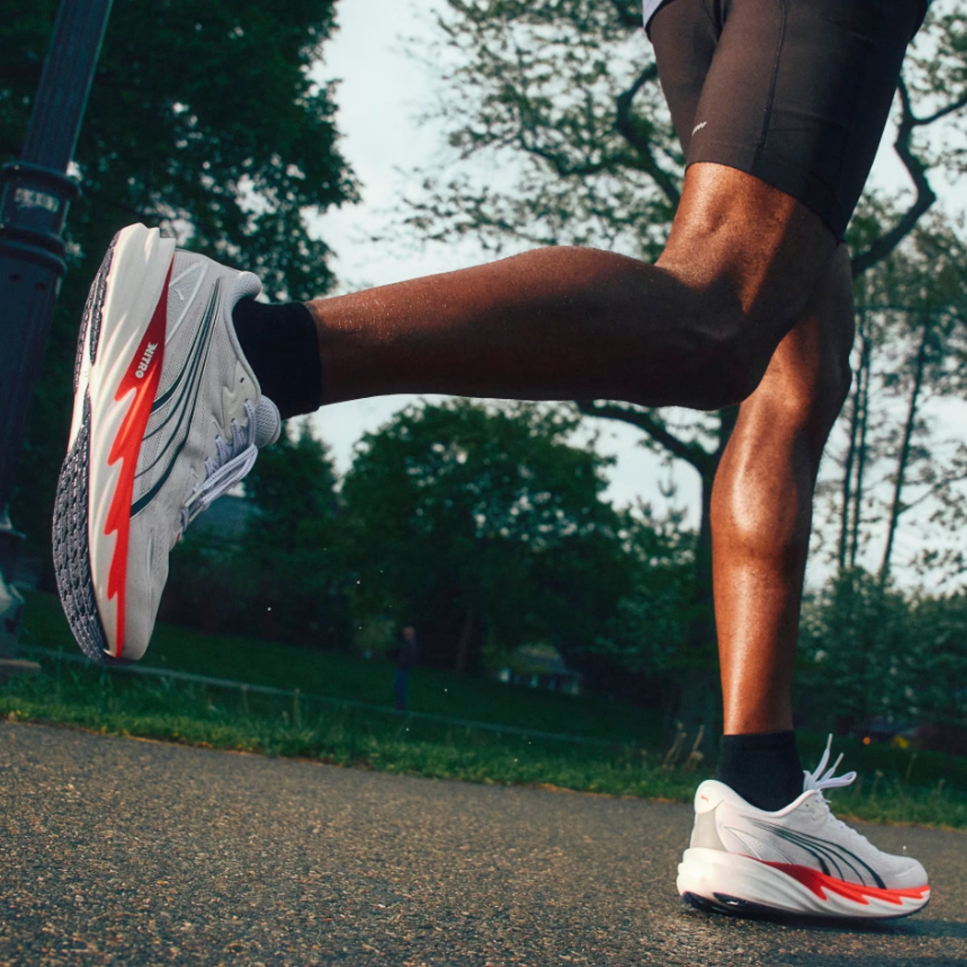 A close-up action shot of a runner's legs in a park, wearing white running shoes with red and gray accents, black socks, and dark running shorts.