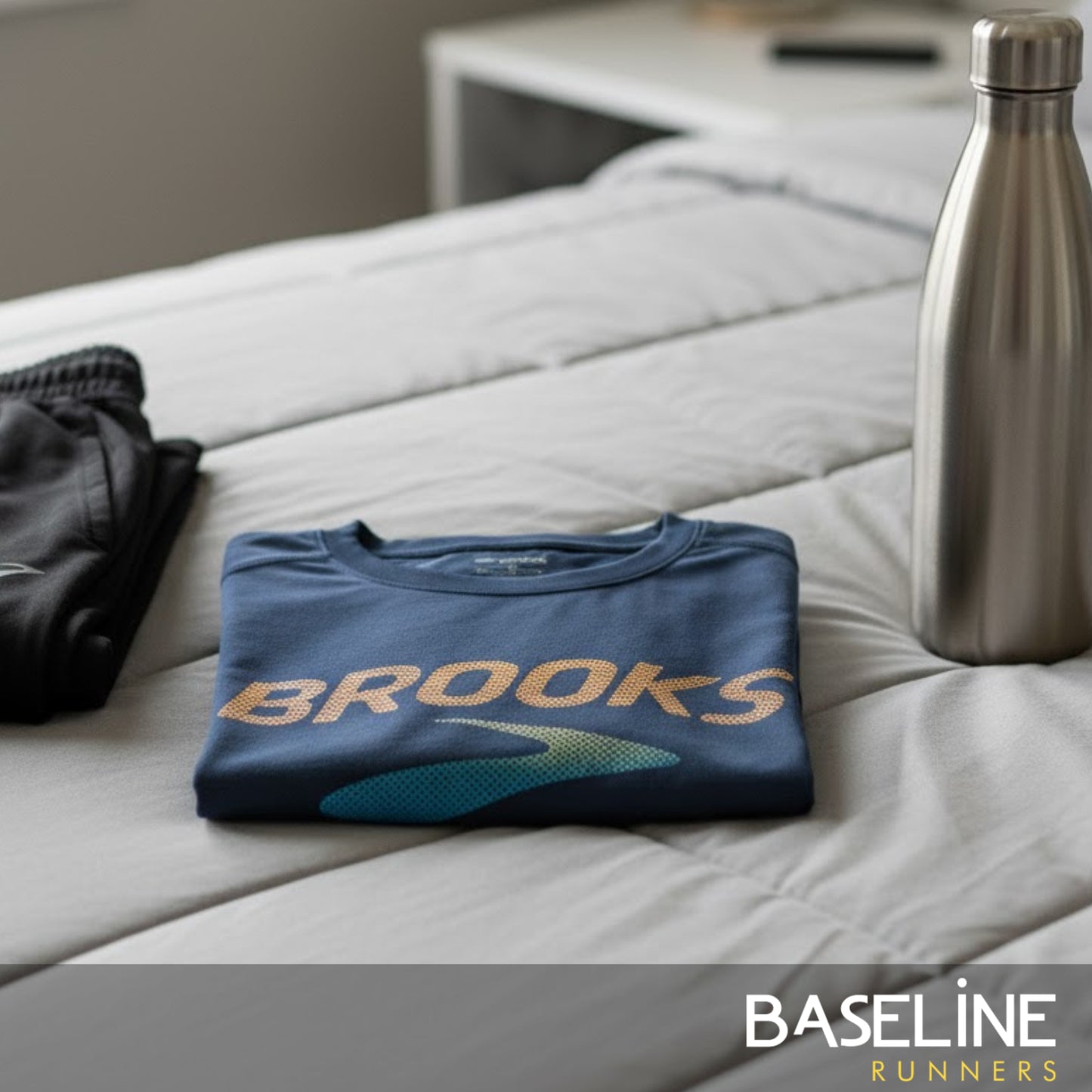 Folded blue shirt with 'Brooks' logo on a bed next to a water bottle, with 'Baseline Runners' branding.