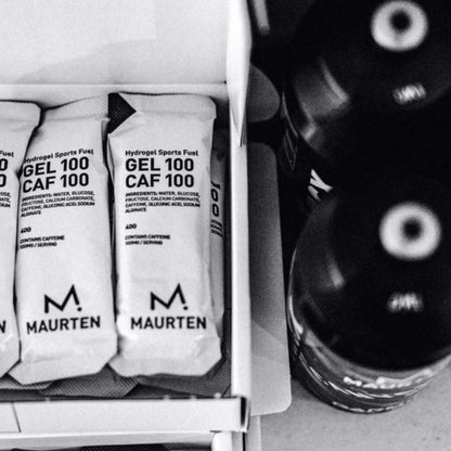 Maurten Hydrogel Sports Fuel gel packets in a white box next to a black bottle, shown in a black and white close-up shot.