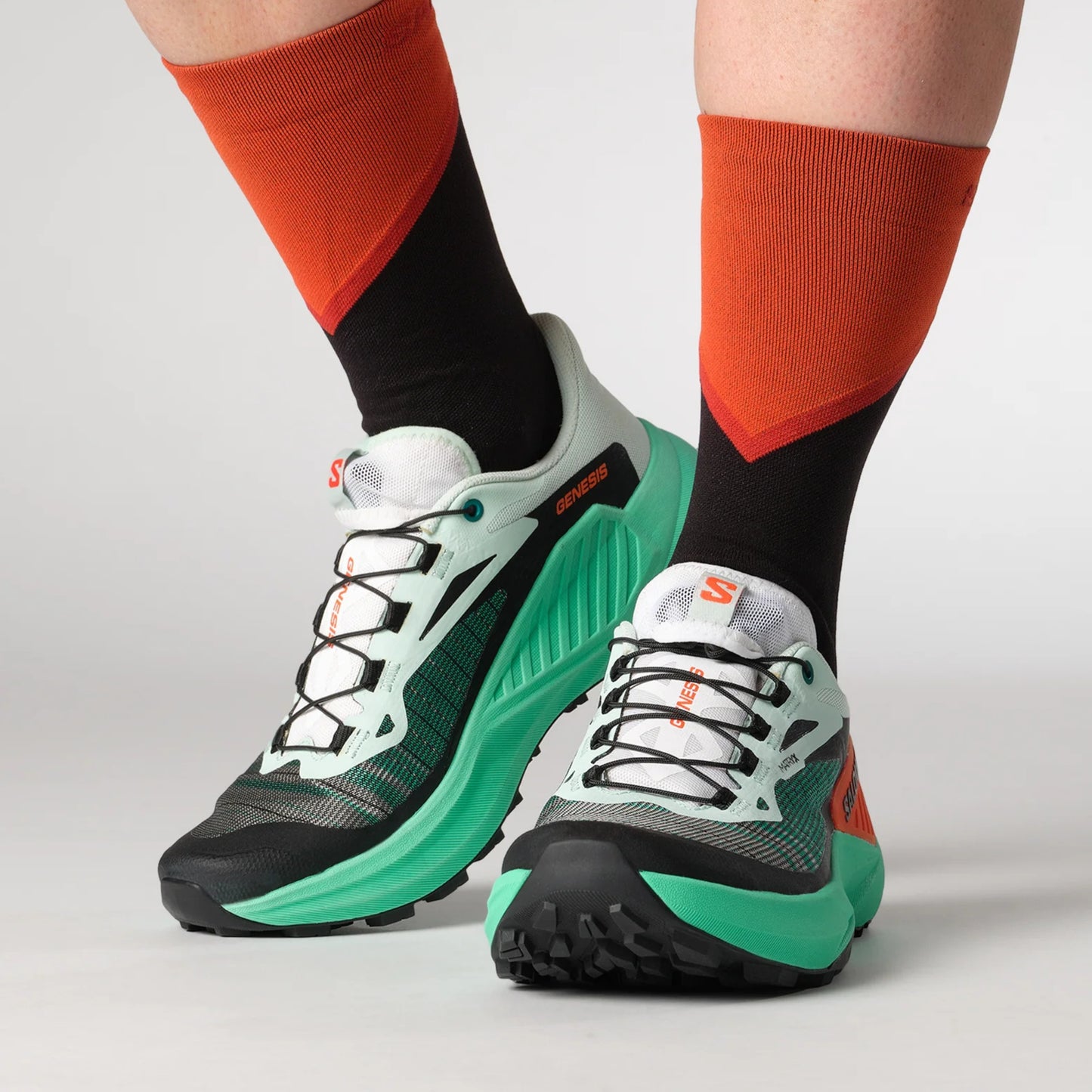 Green and black running shoes with orange accents worn with orange and black socks on a light gray background